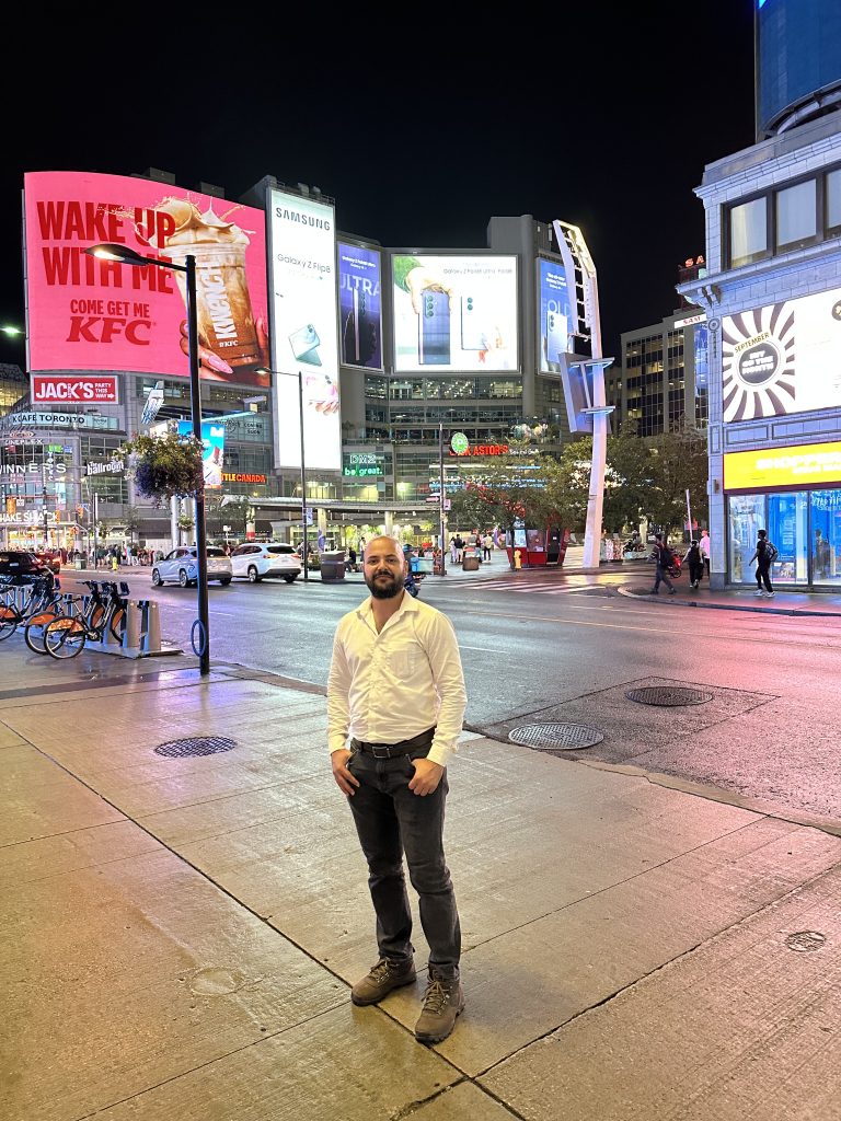 Baljinder Singh standing in a Toronto square surrounded by illuminated signs.